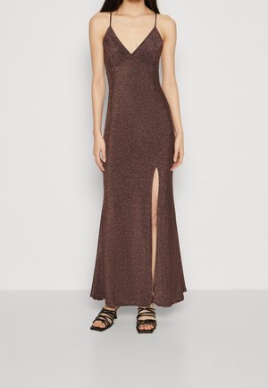 Femme portant une robe maxi marron scintillante sans manches avec une fente haute sur la cuisse et des sandales noires à lanières, debout devant un fond uni.