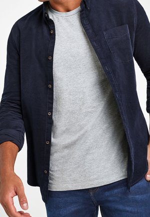 Homme portant un t-shirt gris clair sous une chemise bleu marine foncée déboutonnée avec les manches retroussées et un jean bleu.