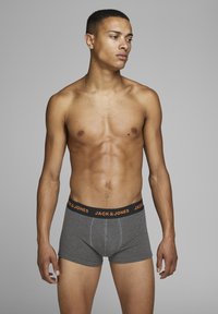 Graue Boxer-Shorts mit einem schwarzen Bündchen, das orangefarbigen Text "JACK & JONES" trägt. Glattes Material, enge Passform und moderner Schnitt.