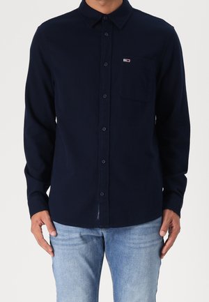 Homme portant une chemise bleu marine à manches longues avec une poche poitrine et un jean bleu clair, debout devant un fond uni.