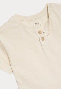 Světle béžové bavlněné tričko s krátkým rukávem, které má Henley výstřih a dvě hnědé knoflíky na léze. Měkká textura, jednoduchý design.