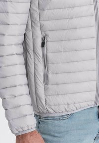 Lys grå quiltet jakke med vandret syninger, lynlåsende sidelomme og elastiske manchetter, lavet af et glat, letvægtsmateriale.