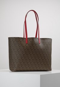 Brun tote-väska med ett upprepande geometriskt mönster, röda läderremmar och en strukturerad form. Materialet har en texturerad yta.