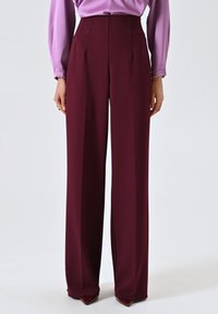 Pantaloni a vita alta e gamba larga in tessuto bordeaux profondo. Caratterizzati da pieghe frontali e una trama liscia. Abbinati a una blusa lilla.