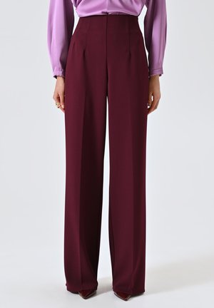 Pantaloni a vita alta e gamba larga in tessuto bordeaux profondo. Caratterizzati da pieghe frontali e una trama liscia. Abbinati a una blusa lilla.