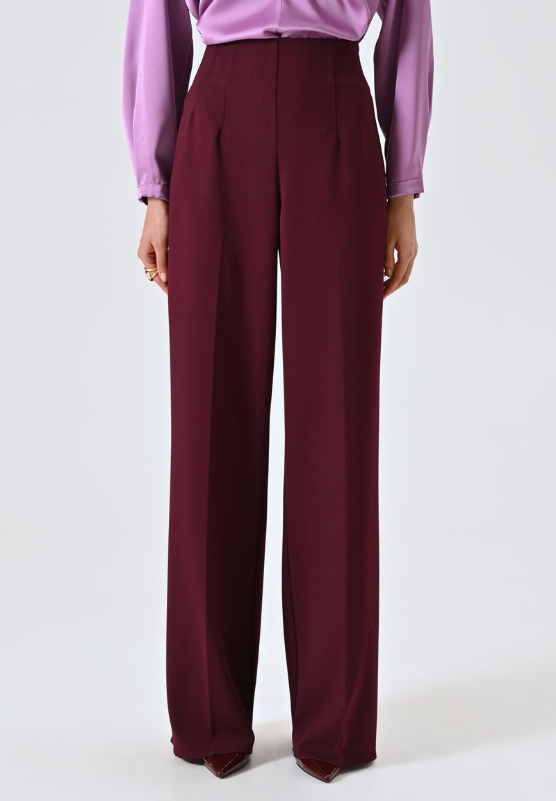 Pantaloni a vita alta e gamba larga in tessuto bordeaux profondo. Caratterizzati da pieghe frontali e una trama liscia. Abbinati a una blusa lilla.