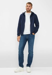 Marineblauer Reißverschluss-Hoodie mit grauem Innenfutter, weißem T-Shirt, blauen Jeans und weißen Sneakern. Der Hoodie hat eine lockere Passform und gerippte Bündchen.