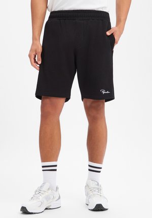 Mann trägt schwarze, gerippte Shorts mit elastischem Bund und weißem Schriftzug-Logo, weiße Sportsocken mit schwarzen Streifen und weiße Sneakers.