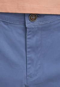 Modré látkové šortky s hladkou texturou, s jedním hnědým knoflíkem a viditelným švem. Částečně zakryté broskvově zbarvenou košilí nahoře.