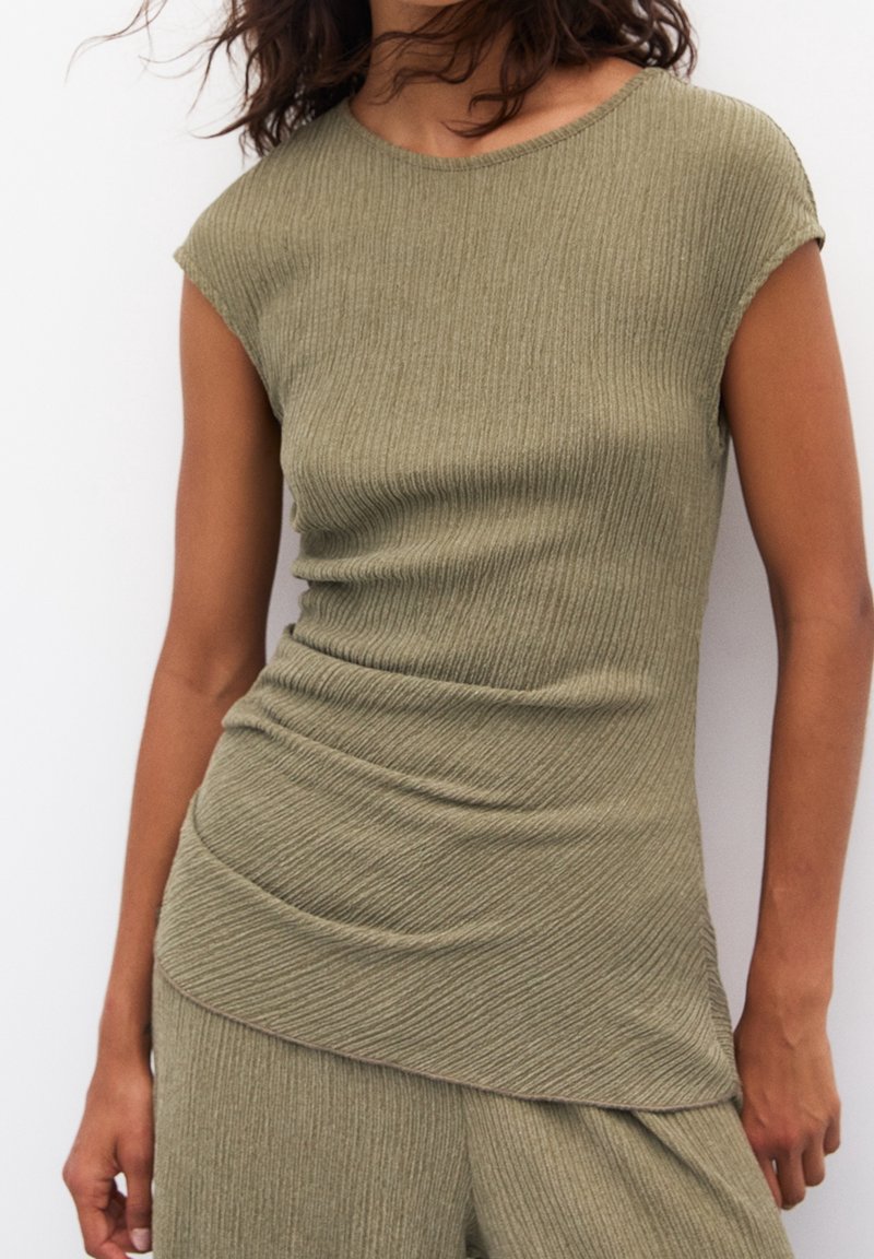 Femme portant un haut court en maille côtelée vert olive avec un ourlet asymétrique et un pantalon côtelé assorti sur un fond uni.
