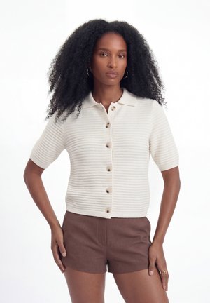 Kvinde med krøllet hår iført en cremefarvet ribbet kortærmet knap-bluse og brune shorts, står med hænderne på hofterne.