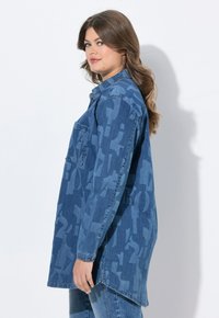 Camicia di denim in varie tonalità di blu, caratterizzata da un audace motivo astratto. Maniche lunghe, polsini con bottoni e taschino sul petto.