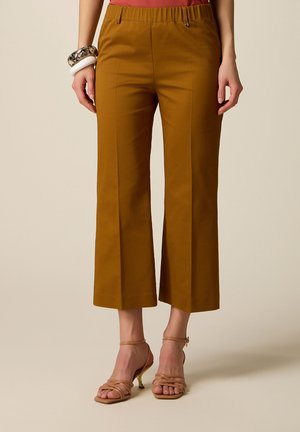 Kvinde iført sennepsfarvede, vidde bukser med elastik i taljen og nøgenfarvede, snørede sandaler med hæl, stående foran en ensfarvet baggrund.