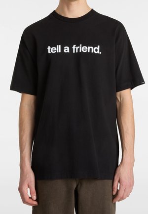 Persoon draagt een zwart T-shirt met witte tekst "tell a friend." en staat voor een effen lichte achtergrond.