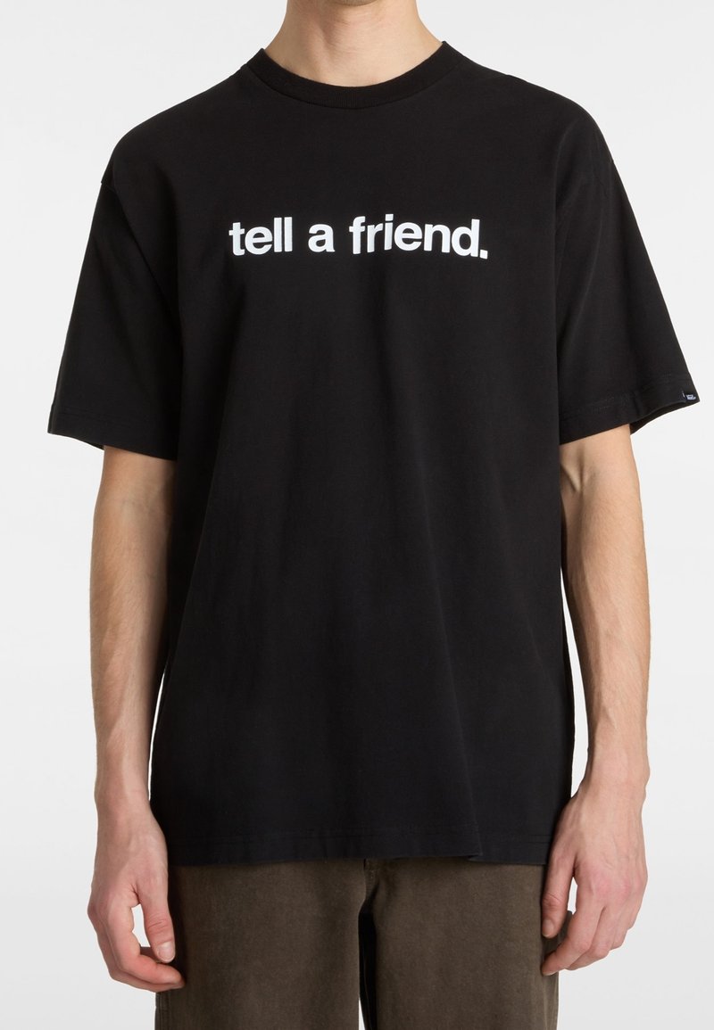 Personne portant un t-shirt noir avec le texte blanc « tell a friend. » debout devant un fond clair uni.