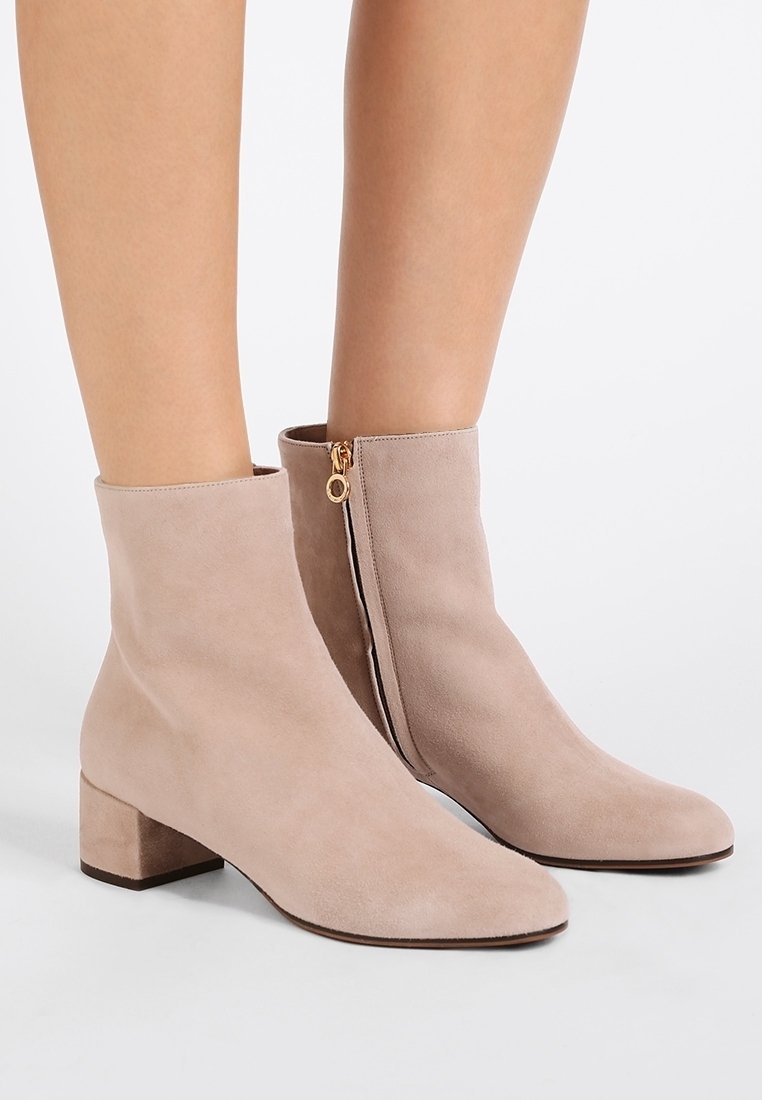 Botas de ante hasta el tobillo en beige claro con punta afilada y tacón bajo en bloque. La cremallera lateral con herrajes dorados añade detalle. Textura suave.