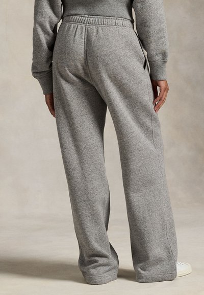 Άτομο που φοράει γκρι φαρδιά αθλητική φόρμα και αντίστοιχο φούτερ, στέκεται με το χέρι ακουμπισμένο στο πλάι, μπεζ φόντο.