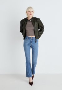 Jachetă din piele verde măsliniu cu guler înalt, asortată cu un top scurt maro și blugi deschiși la culoare cu evazare. Pantofii cu toc ascuțit burgund finalizează look-ul.
