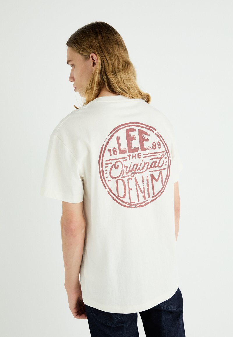 Cremefarbener Baumwoll-T-Shirt mit einem großen roten Drucklogo auf der Rückseite, das "LEE 1889 The Original DENIM" lautet. Kurze Ärmel, lockere Passform.