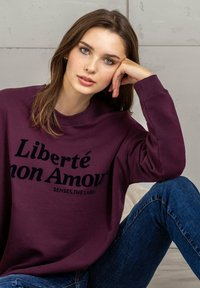 Maroon sweatshirt with black text "Liberté mon Amour" in bold font, featuring a relaxed fit and ribbed neckline, paired with blue denim jeans.