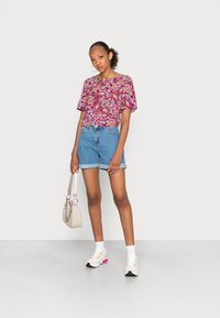 Camisa de manga curta com padrão floral em bordô, combinada com calções de ganga. Os acessórios incluem uma pequena mala clara e ténis coloridos.