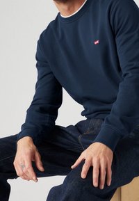 Mörkblå sweatshirt i bomullsblandning, med ribbad rund hals och muddar. Litet rött Levi's-logotyp på bröstet, tillsammans med mörka denimjeans.