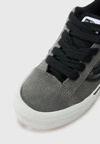 Gray suede sneaker with black laces, featuring a textured surface and a white rubber sole. Notable design includes a side logo accent.