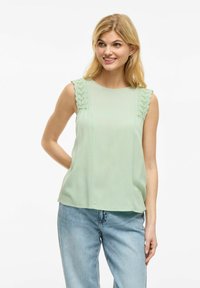 Top sin mangas de color verde claro con acentos de encaje texturizado en los hombros, corte relajado, escote redondo y tela suave. Combinado con jeans azules.