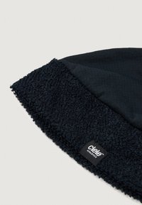 Căciulă neagră dintr-un material fin, cu o margine căptușită cu fleece. Materialul principal este neted, în timp ce marginea are o textură pufoasă. Eticheta brandului este vizibilă.