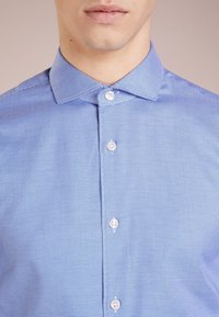 Camicia a quadri blu con colletto classico, sei bottoni bianchi e un motivo di tessuto strutturato.