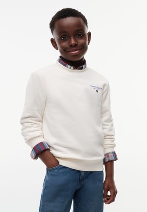 Junge lächelt mit den Händen in den Taschen, trägt ein weißes Tommy Hilfiger Sweatshirt über einem rot-blau karierten Hemd und blaue Jeans.