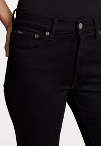 Černé džíny z denimu s úzkým střihem, disponující standardními kapsami, zipem a jemnou značkovou nášivkou. Hladká textura s minimálními detaily.