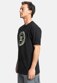 Černé bavlněné tričko s krátkým rukávem, se zaobleným grafikou na bílé barvě, zobrazující logo DC a text v kontrastním provedení.