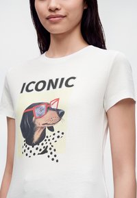 Jeune femme porte un t-shirt blanc avec une illustration d'un teckel portant des lunettes rouges et des pois noirs, le texte au-dessus dit "ICONIC".