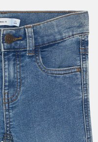 Jeans in denim azzurro chiaro con zip, chiusura con bottone e cinque tasche. Le cuciture sono di un colore dorato a contrasto, con leggere sfumature.