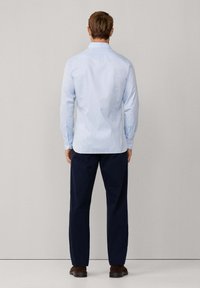 Man met de rug naar de kijker, gekleed in een lichtblauwe geruite overhemd met lange mouwen, donkeren marineblauwe broek en bruine schoenen, tegen een effen achtergrond.