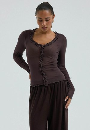 Femme portant un haut ajusté à manches longues marron foncé avec bordure à volants et un pantalon marron foncé ample, debout devant un fond clair uni.