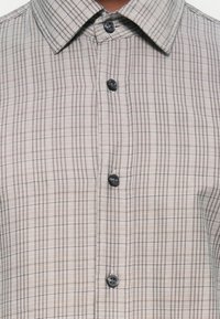 Knappskjorte med et lysebeige og mørkebrunt rutemønster, spredt krage, stoffet har en glatt tekstur med en subtil glans.