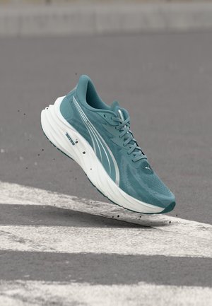 Teal running shoe with white sole and laces, mid-air above asphalt road with white markings, small debris beneath the shoe.