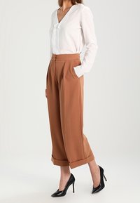 Chemise blanche à manches longues avec un col en V, associée à un pantalon large marron avec des poches latérales et un ourlet court, portée avec des talons noirs.