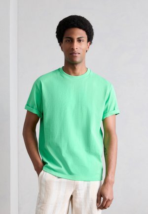 Άνδρας στέκεται απέναντι σε απλό τοίχο, φορώντας πράσινο μπλουζάκι με γυρισμένα μανίκια και μπεζ ριγέ παντελόνι, με το ένα χέρι στην τσέπη.