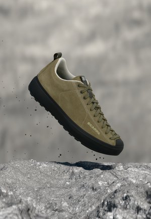 Scarpa da ginnastica verde oliva con suola e lacci neri che fluttua sopra un terreno roccioso, mostra la vista laterale e il nome del marchio "Scarpa".