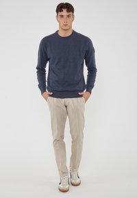 Maglione a maglia blu navy con scollo rotondo, chinos beige e sneakers bianche con dettagli blu. Il modello è in piedi con le mani nelle tasche.