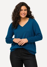 Teal gestrickter Pullover mit V-Ausschnitt, langen Ärmeln und gerippten Bündchen. Kombiniert mit dunklen Jeans, die einen lockeren Schnitt und eine glatte Textur aufweisen.