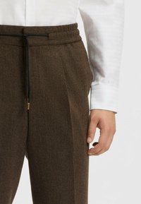 Bruine herringbone trousers van een gestructureerde stof, met een aansluitende tailleband en zwarte trekkoord. G gedragen met een wit shirt.