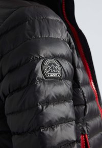 Veste matelassée noire avec un design en coussins et une fermeture éclair rouge. Comprend un écusson rond étiqueté "JACKIT EXPEDITION ALPS DOWN."