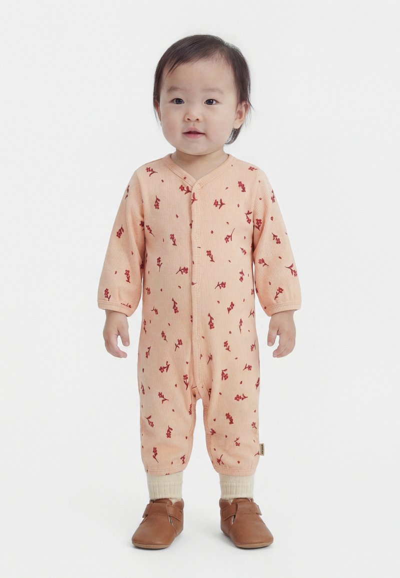 Kisgyermek áll, barackszínű hosszú ujjú napozóruhát visel apró piros virágmintákkal, krémszínű zoknit és barna cipőt, előre néz fehér háttér előtt.