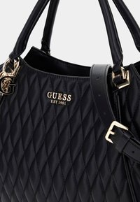 Zwarte gewatteerde leren Guess handtas met gouden logo, gouden gesp en dubbele handvatten tegen een witte achtergrond.