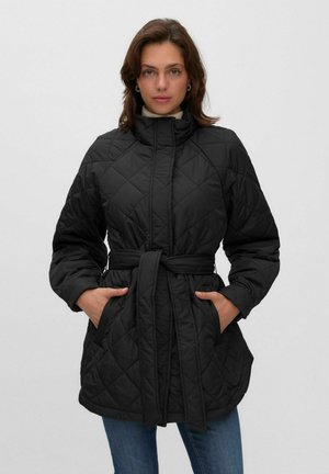 Frau trägt eine schwarze gesteppte Jacke mit hohem Kragen und gebundenem Gürtel, Hände in den Taschen, steht vor einem schlichten hellen Hintergrund.