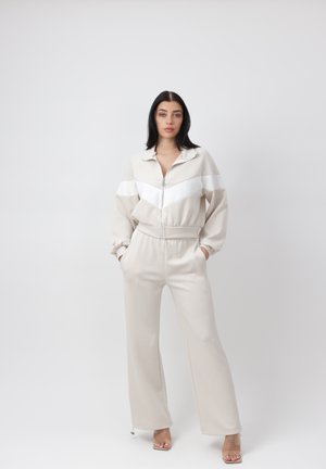 Kobieta stojąca z rękami w kieszeniach, ubrana w beżowo-białą kurtkę na zamek i pasujące szerokie spodnie oraz przezroczyste sandały na obcasie.